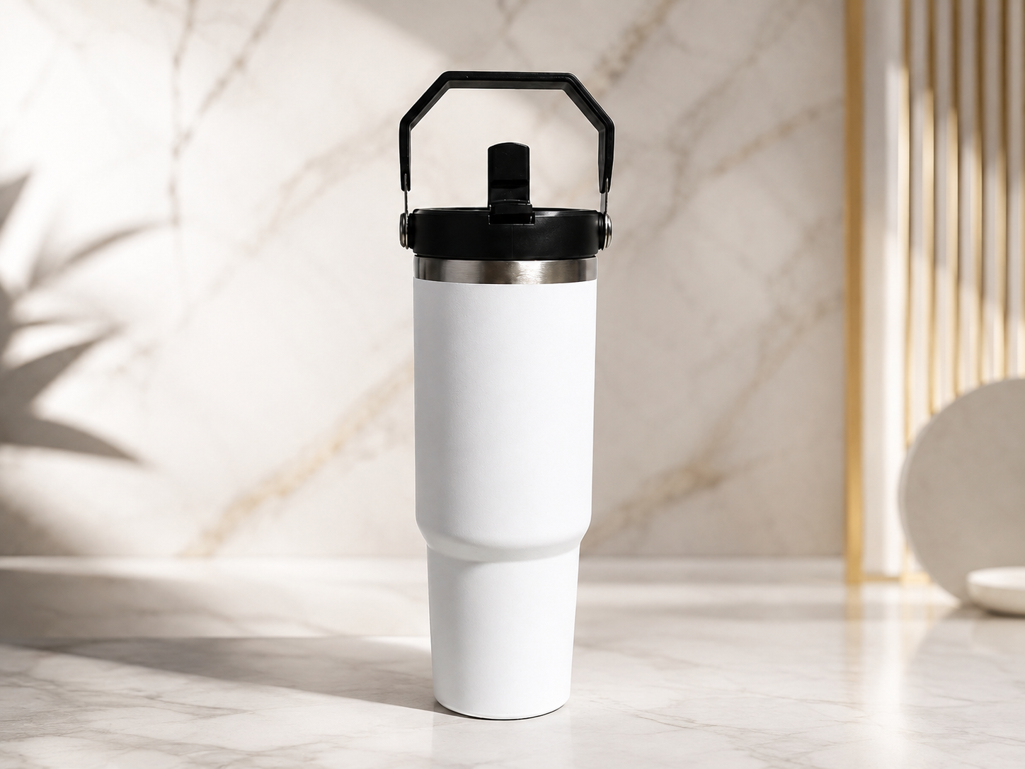 A white insulated travel tumbler with a black handle lid, standing on a marble surface against a marble wall background.