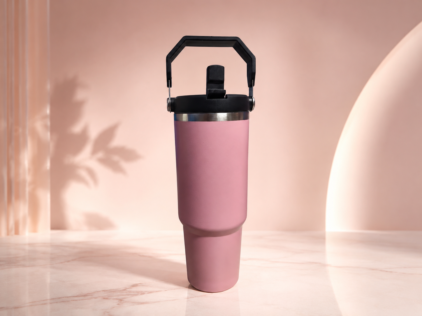 A matte pink insulated travel tumbler with a black handle lid and a silver metal band, standing on a light marble surface against a warm beige background with soft shadows.