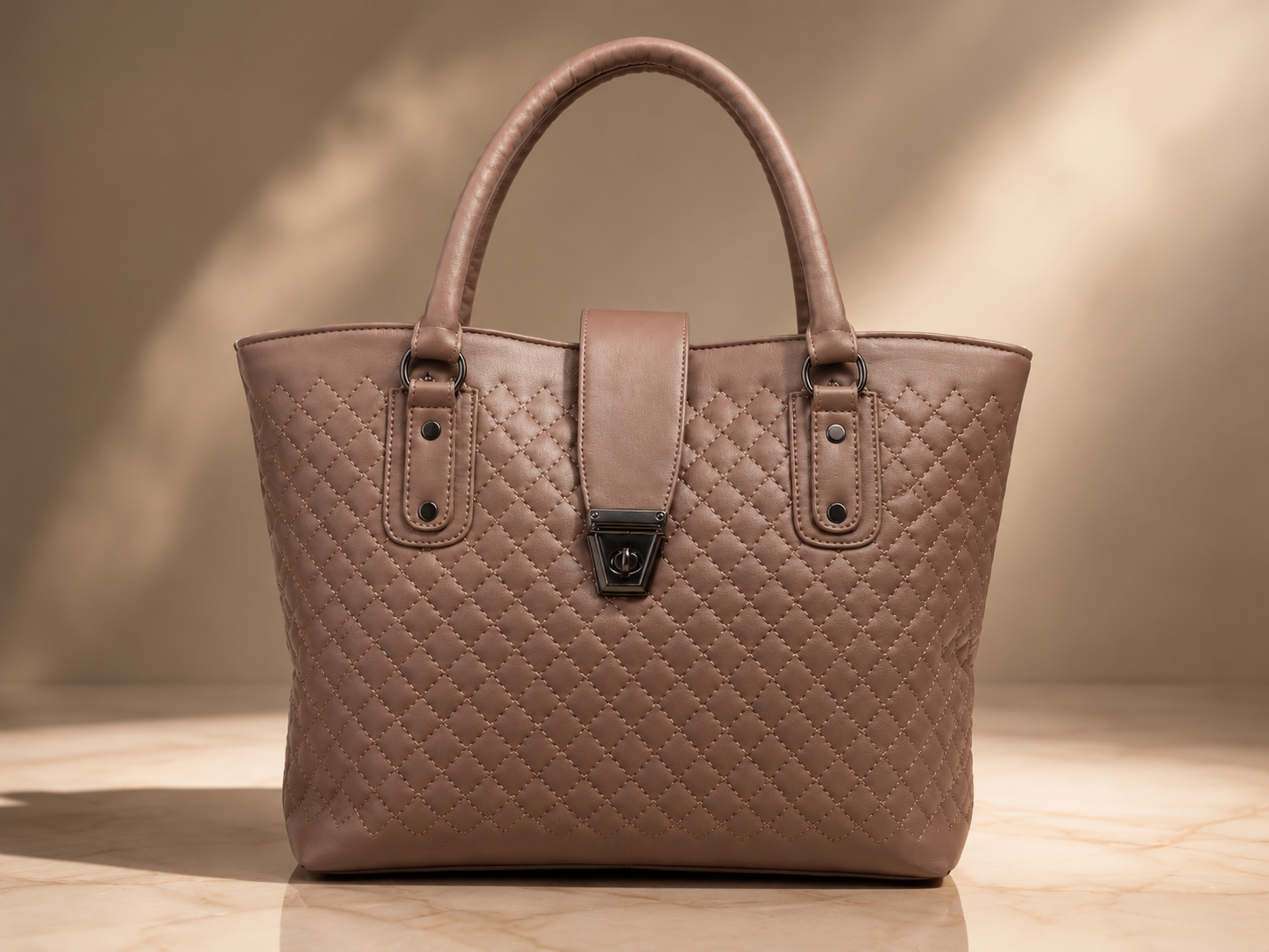 A structured quilted leather tote bag in a warm taupe-brown color, featuring dual top handles, a central strap closure with a black metal clasp, and a diamond-quilted pattern, set against a warm beige background with soft shadows.